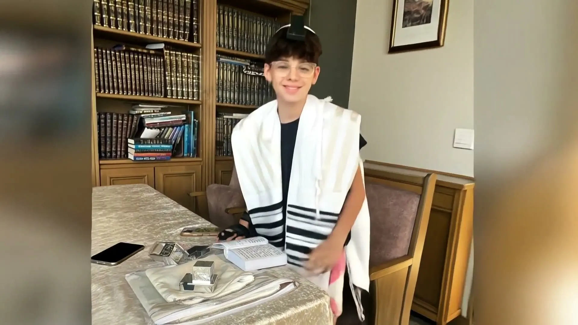 פריים מתוך הסרטון — חתן בר המצווה עטוף בטלית, מחייך בחדר עם ספרייה